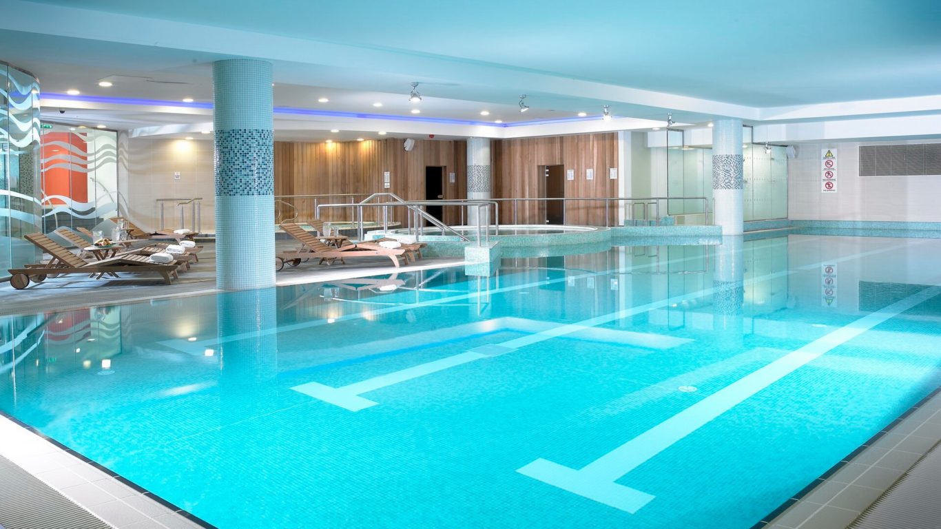 Indoor swimming pool with clear blue water, lounge chairs, and towel racks along the side; wood paneling and glass doors in the background.