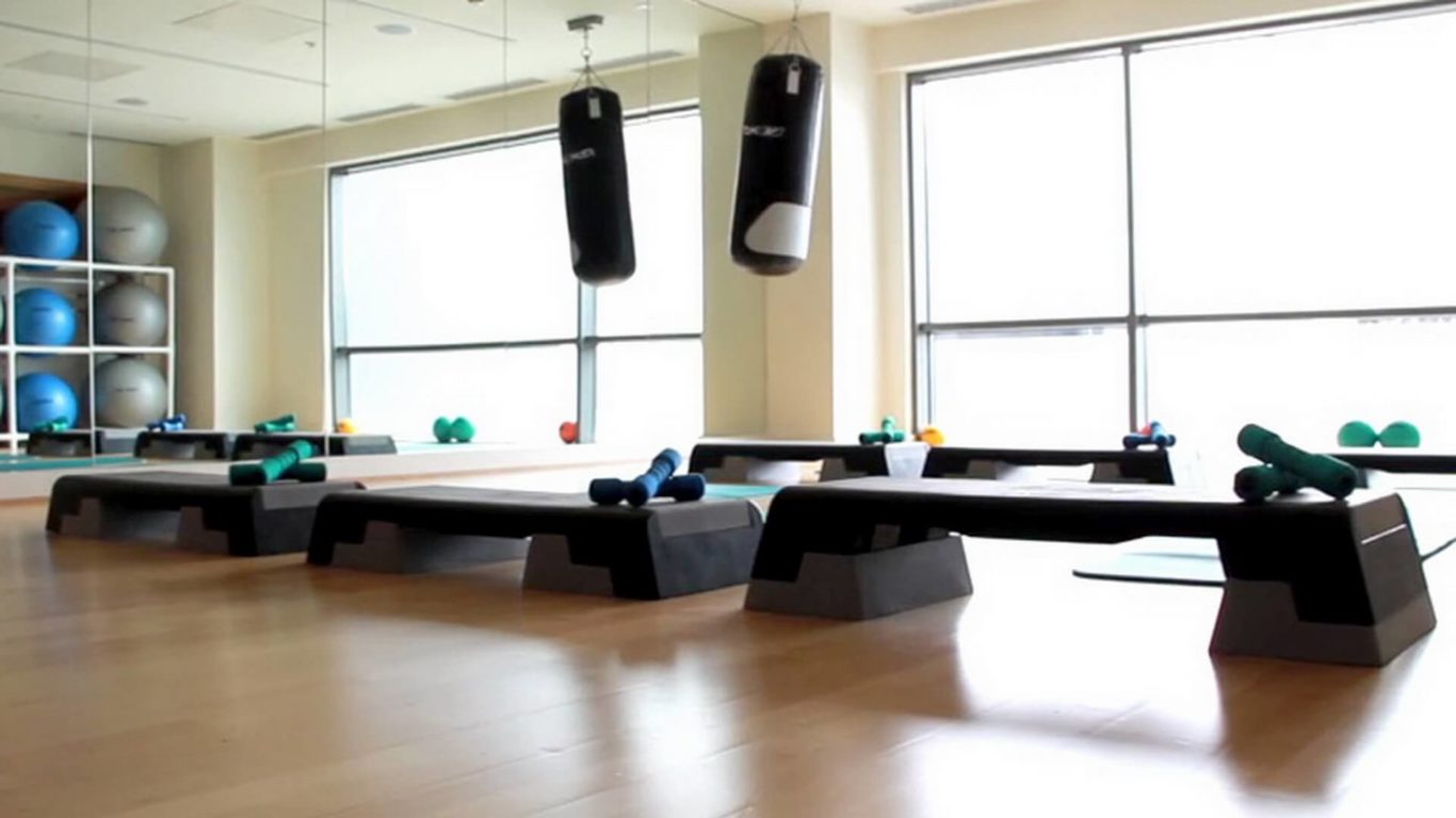 A fitness studio with step platforms, hand weights, yoga mats, exercise balls, and two punching bags hanging from the ceiling, next to a wall of mirrors and large windows.