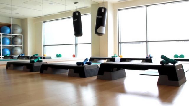 A fitness studio with step platforms, dumbbells, two hanging punching bags, exercise balls on shelves, and large windows letting in natural light.
