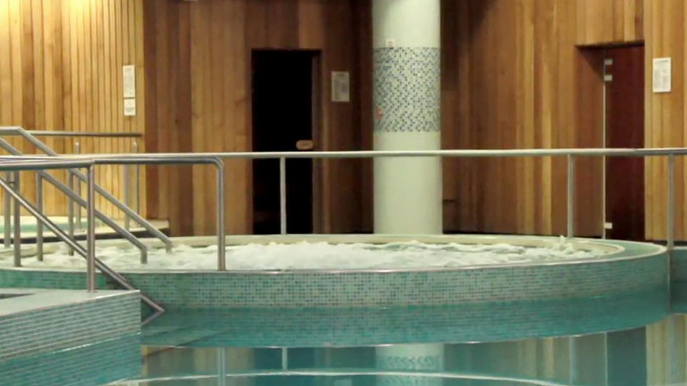 Indoor spa area with a bubbling circular hot tub, metal handrails, tiled pool, and wooden paneled walls.