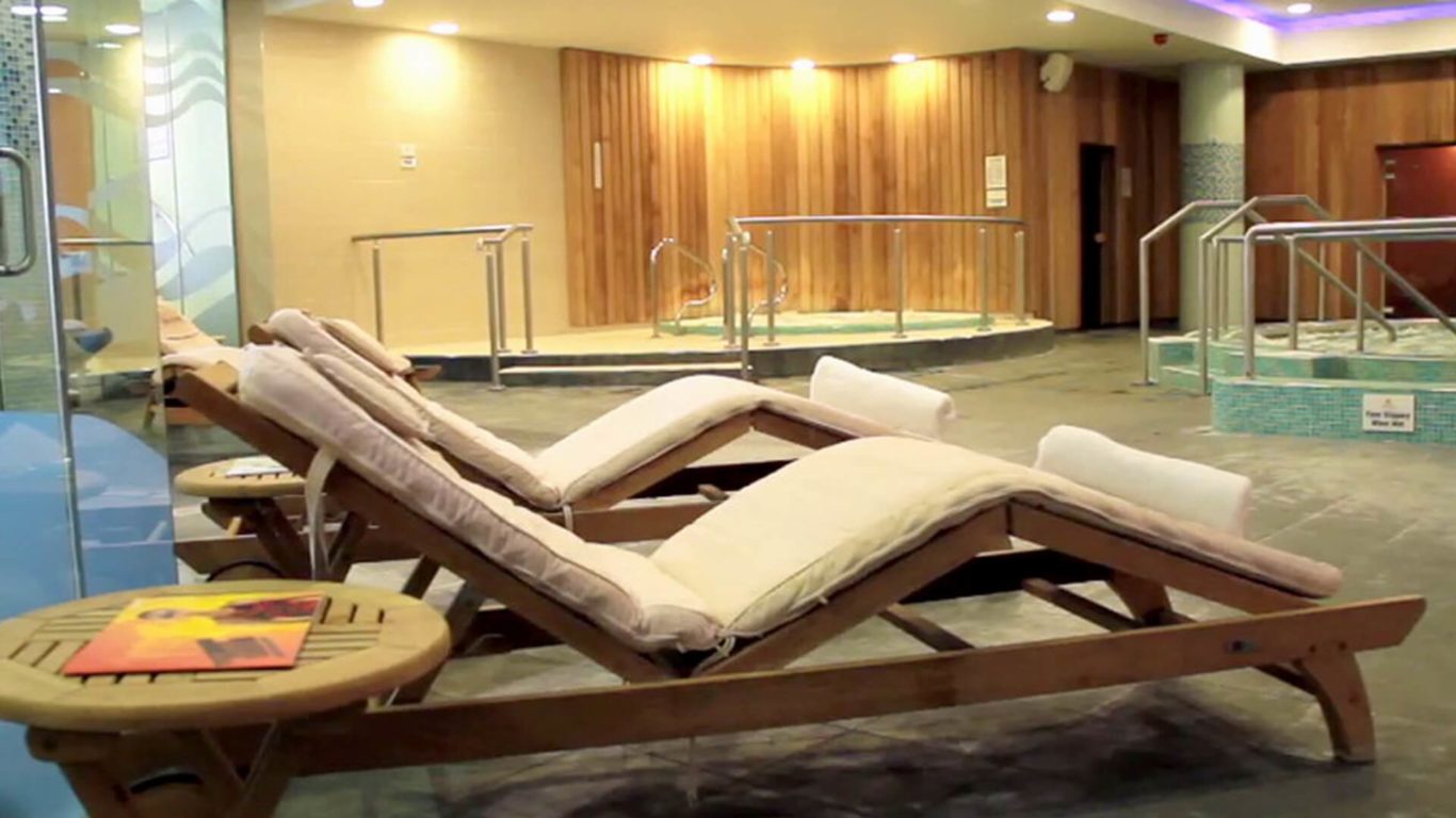 Wooden lounge chairs with cushions are positioned near a round jacuzzi and small pool in a spa relaxation area with wooden panel walls.