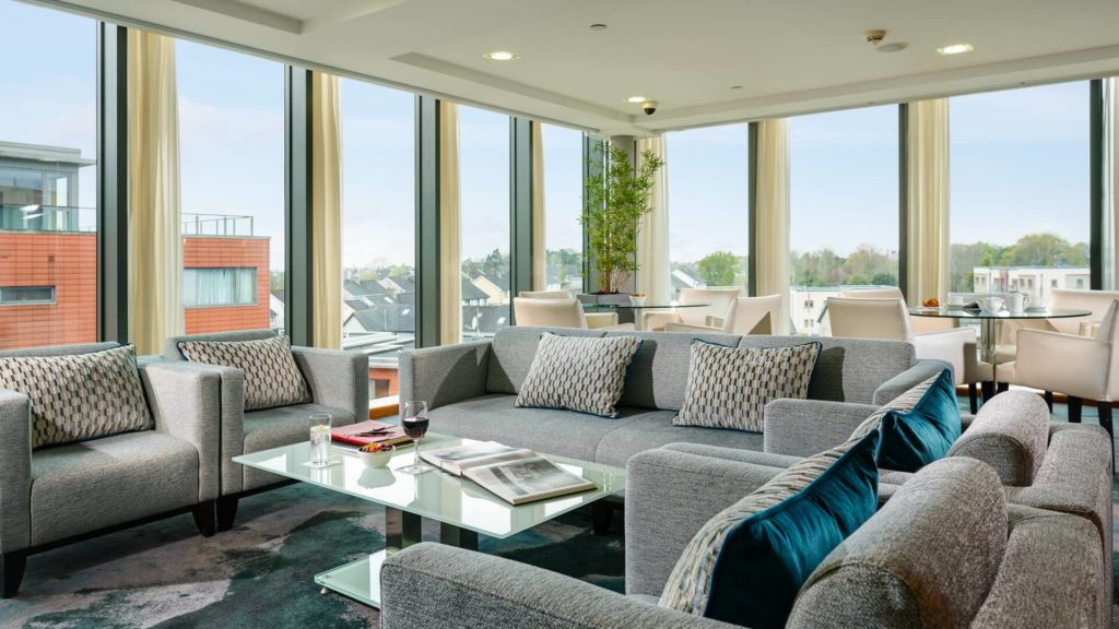 Modern living room with large windows, grey sofas, glass coffee table, and a dining area; cityscape and trees visible outside.