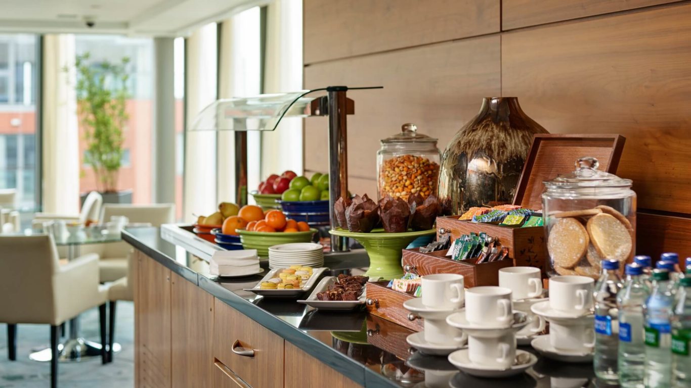 A breakfast buffet with fruit, pastries, muffins, glass jars of snacks, bottled water, stacked teacups, and condiments on a counter in a bright dining area.
