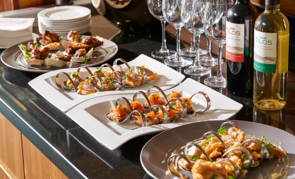 A selection of appetizers arranged on white plates, two bottles of wine, and several empty wine glasses on a black countertop.