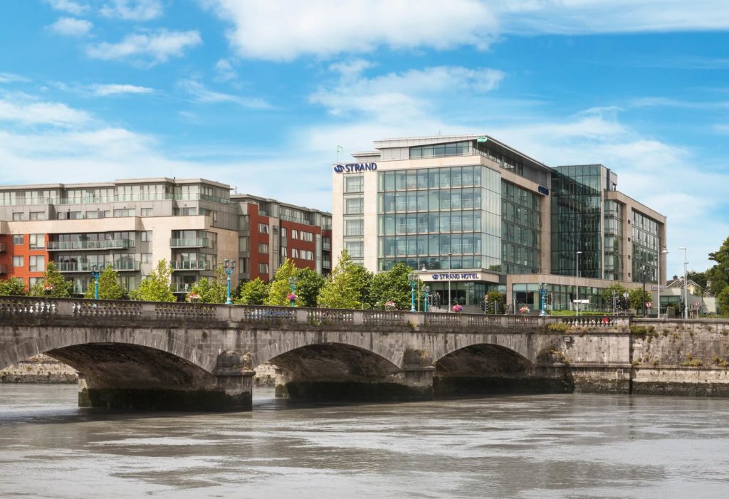 Modern glass and brick buildings, including a hotel, line the riverside behind a stone bridge on a clear day.
