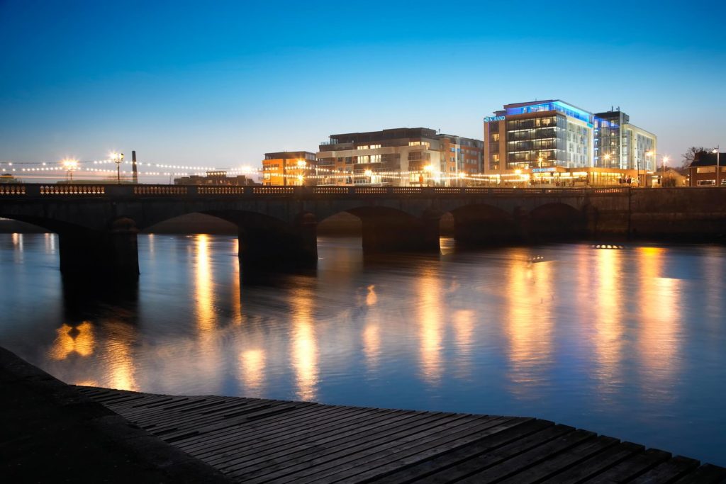 Evening view of a city bridge over a river, with modern buildings and streetlights reflecting on the water under a clear blue sky.