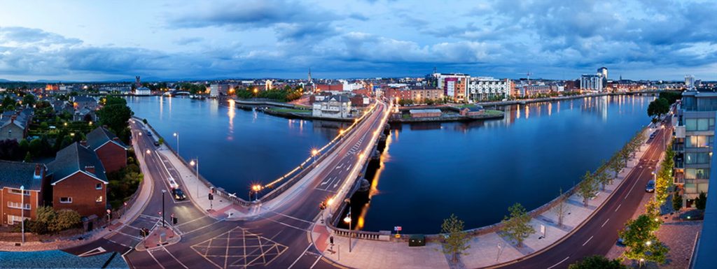 A panoramic view of a cityscape with roads and bridges crossing a wide river, surrounded by buildings under a cloudy sky.