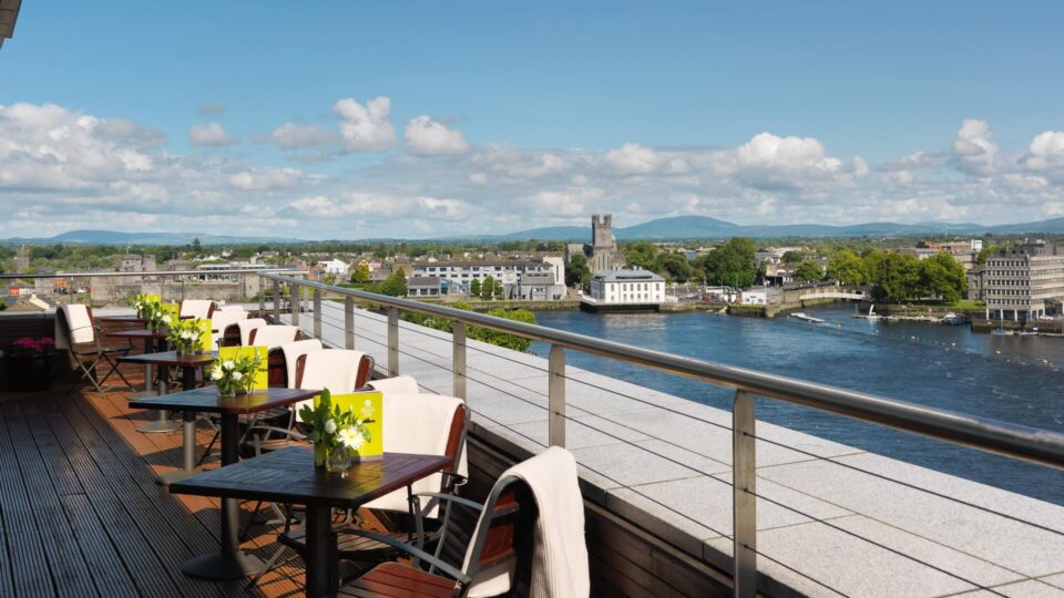 Outdoor terrace with tables and chairs overlooking a river, cityscape, and distant mountains under a blue sky—perfect for relaxing while enjoying exclusive Hotel Deals Limerick.