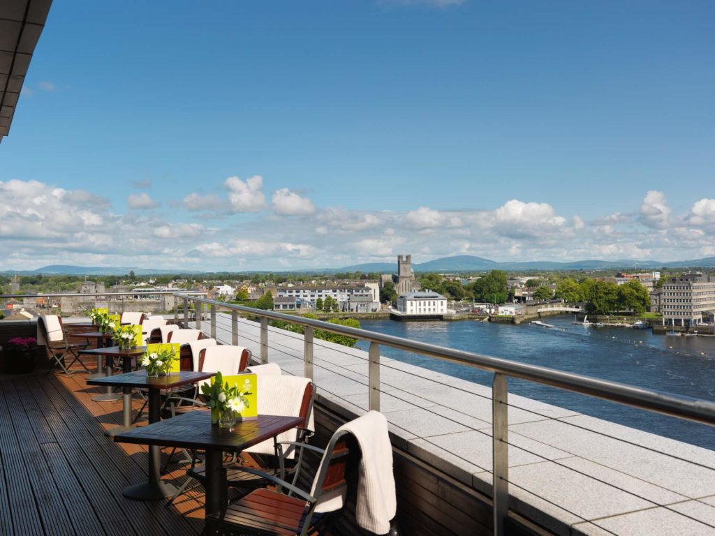 Rooftop terrace with tables and chairs overlooks a river and cityscape under a blue sky with scattered clouds.