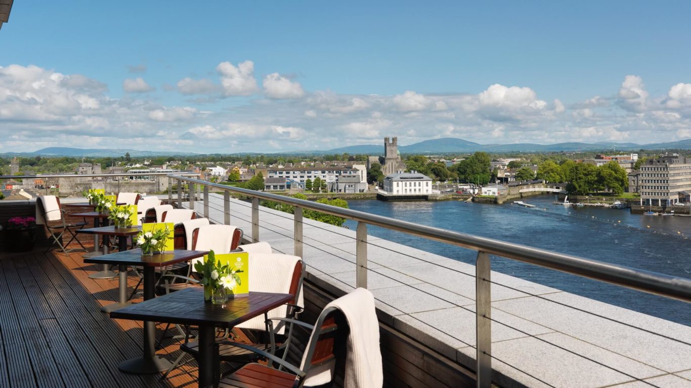 Outdoor terrace with tables and chairs overlooking a river, city buildings, and a distant castle under a partly cloudy sky.