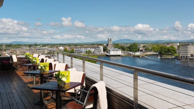 Outdoor terrace with tables and chairs overlooking a river, city buildings, and a distant castle under a partly cloudy sky.