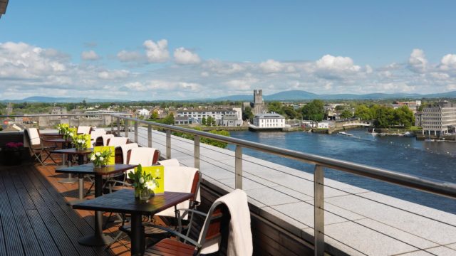 A rooftop terrace with tables and chairs overlooks a river and a cityscape with historic buildings and distant hills under a partly cloudy sky.