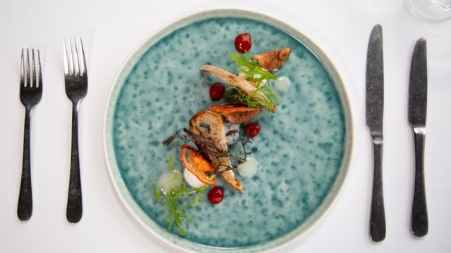 A blue ceramic plate with an artfully arranged dish including roasted vegetables, sauce dots, and microgreens, set on a white tablecloth with a fork and knife on each side.