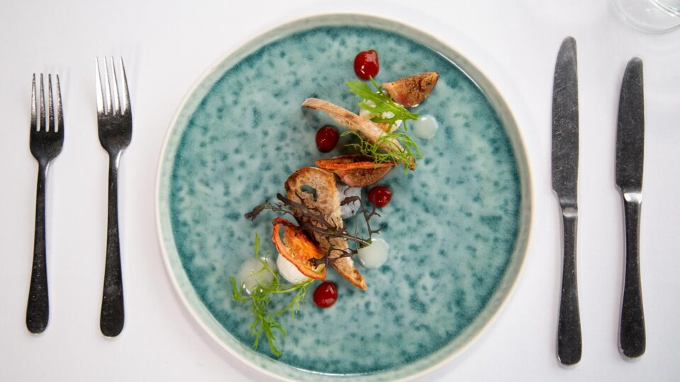 A blue ceramic plate with an artfully arranged dish including roasted vegetables, sauce dots, and microgreens, set on a white tablecloth with a fork and knife on each side.
