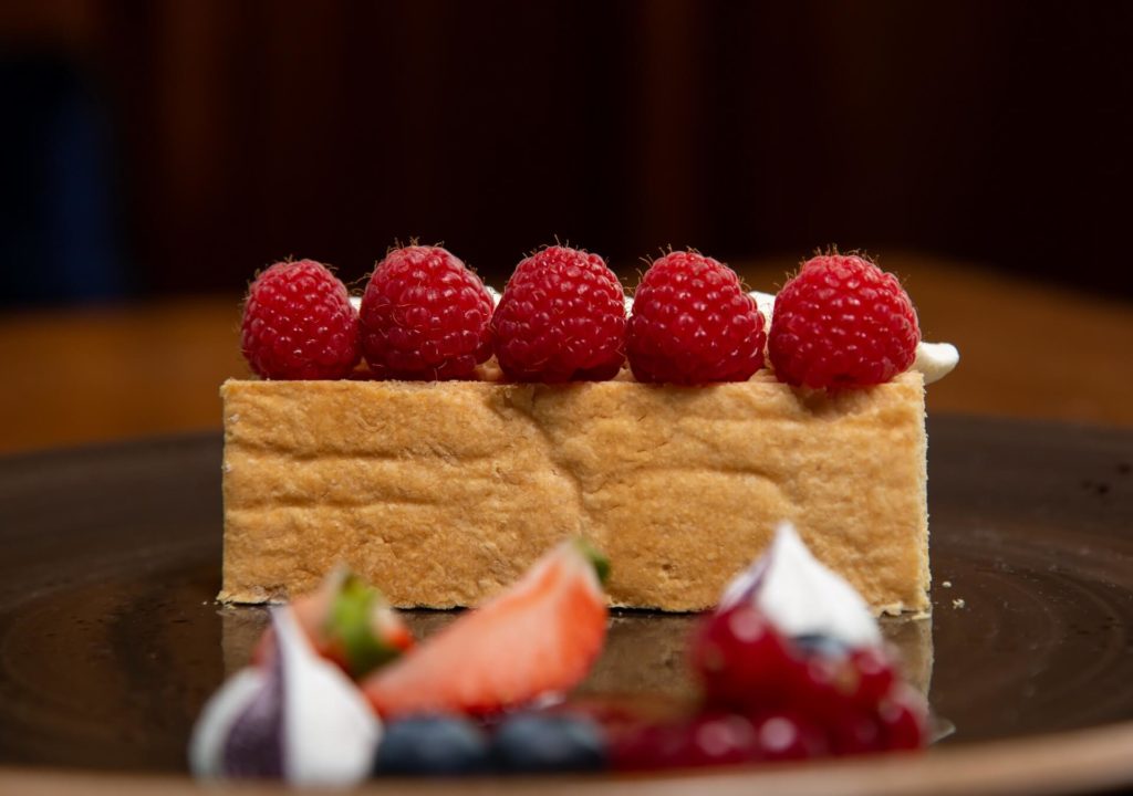 A rectangular slice of layered cake topped with five raspberries sits on a brown plate, with assorted berries and cream garnish in the foreground.