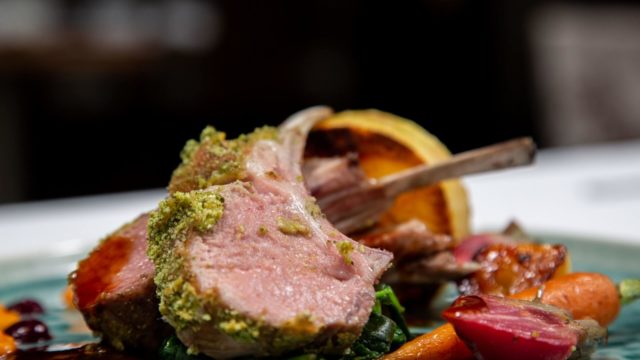 Plated rack of lamb with a green herb crust, served with roasted vegetables and greens on a blue plate.