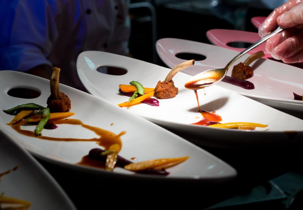 A chef spooning sauce onto a plated gourmet dish with lamb chops, colorful vegetables, and artistic sauce presentation.