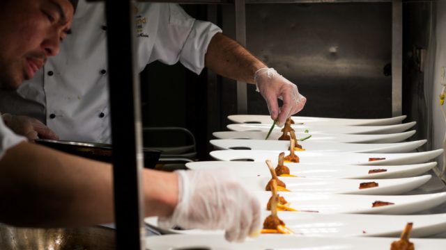 Chefs in a kitchen plating multiple dishes on white plates, carefully arranging food with tongs and gloved hands.
