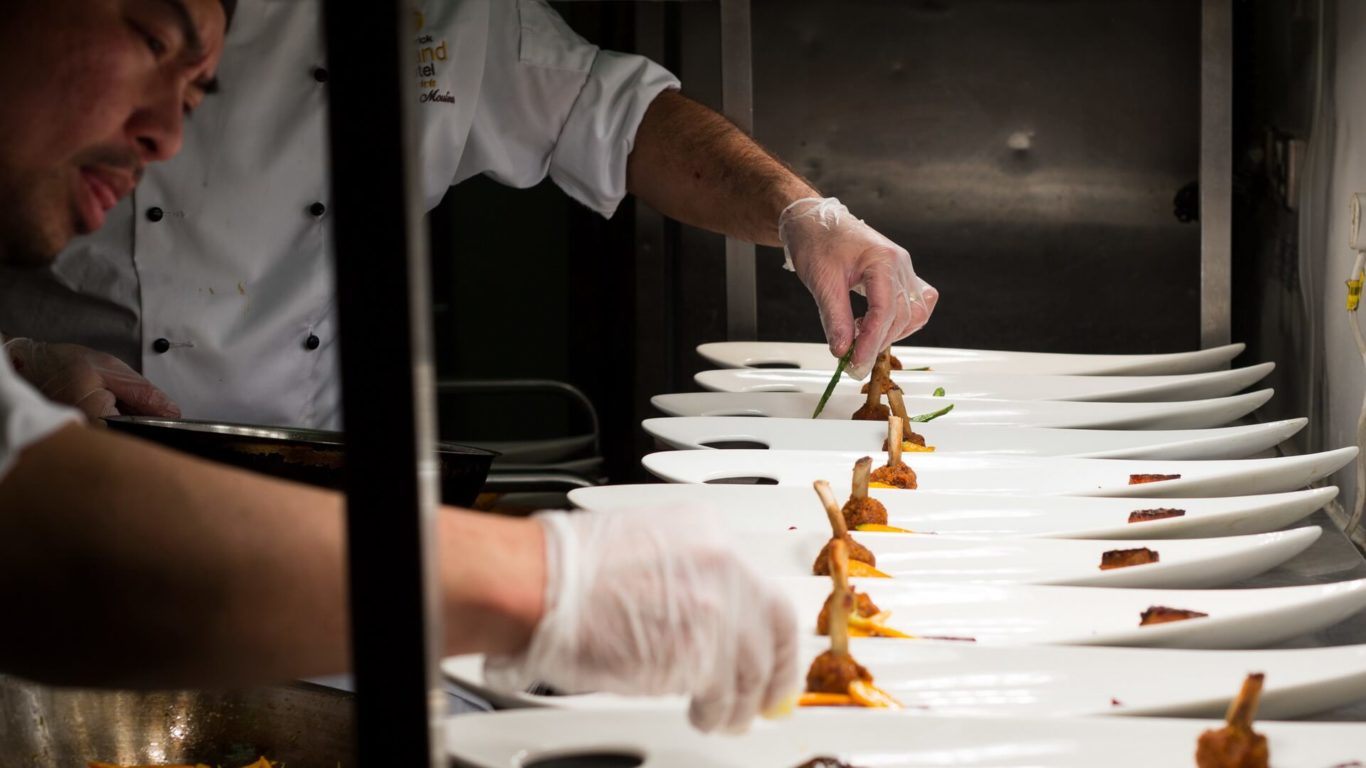Two chefs in white uniforms and gloves carefully plating gourmet food on a row of white dishes in a professional kitchen.