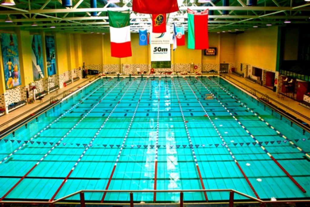 Indoor Olympic-size swimming pool with marked lanes, lane ropes, and flags above. Several national flags hang from the ceiling. Walls are yellow with some banners and artwork.