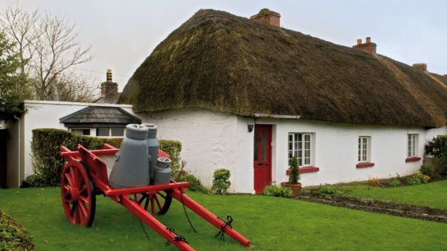 A white cottage with a thatched roof and red door sits behind a red wooden cart holding two gray milk churns on a neatly trimmed green lawn.