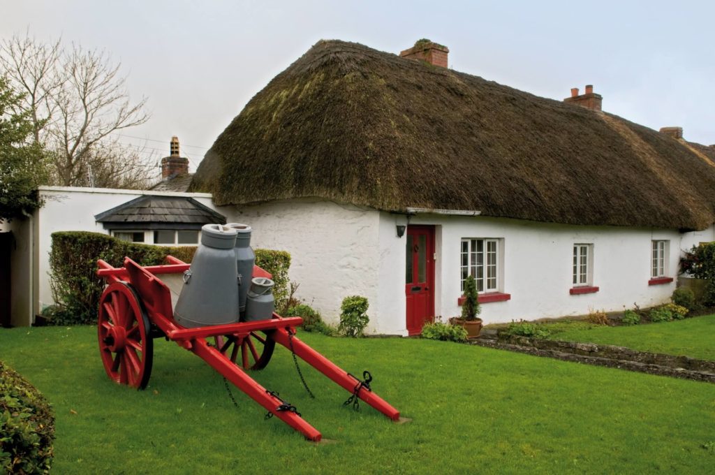 A white thatched cottage with red trim sits behind a lawn, featuring a red wooden cart holding two large milk churns in the garden.