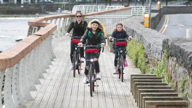 Three people ride bicycles along a wooden riverside path bordered by a stone wall and metal railing on a cloudy day.