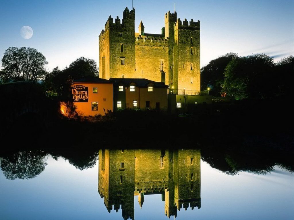 A large stone castle is illuminated at dusk, reflected in a calm body of water, with a full moon visible in the sky and a lit building labeled 