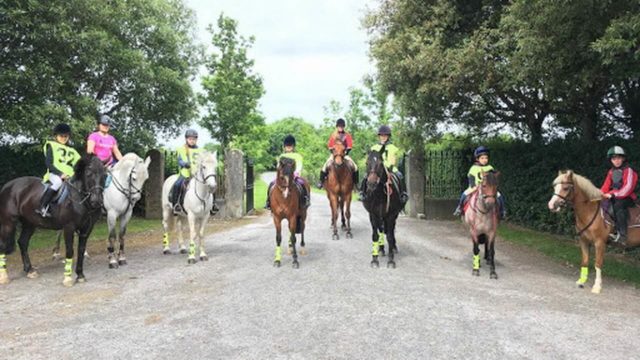 A group of eight children wearing helmets ride horses and ponies on a gravel path, with trees and greenery in the background.
