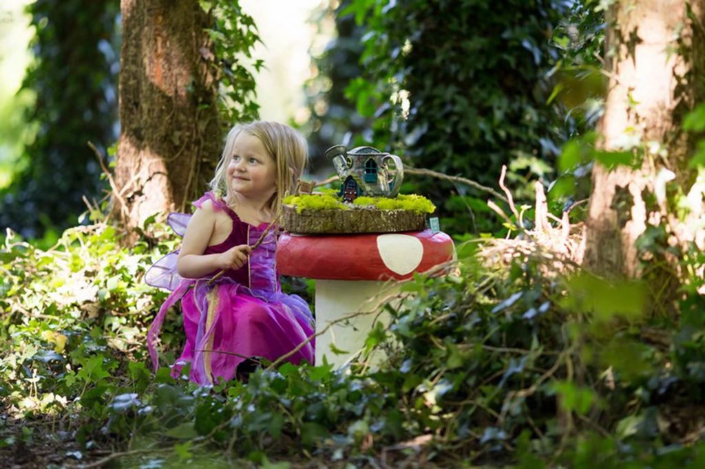 A young girl in a pink fairy costume sits in a forest next to a large mushroom table with a miniature house and teapot set on top.