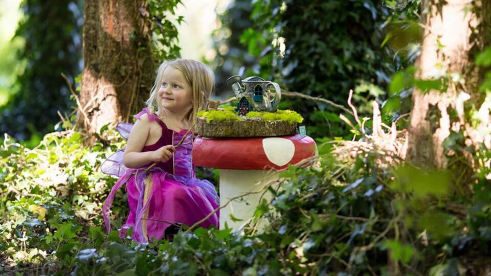 A young girl in a pink dress sits next to a table shaped like a mushroom in a forest, with a small toy house on top of the table.