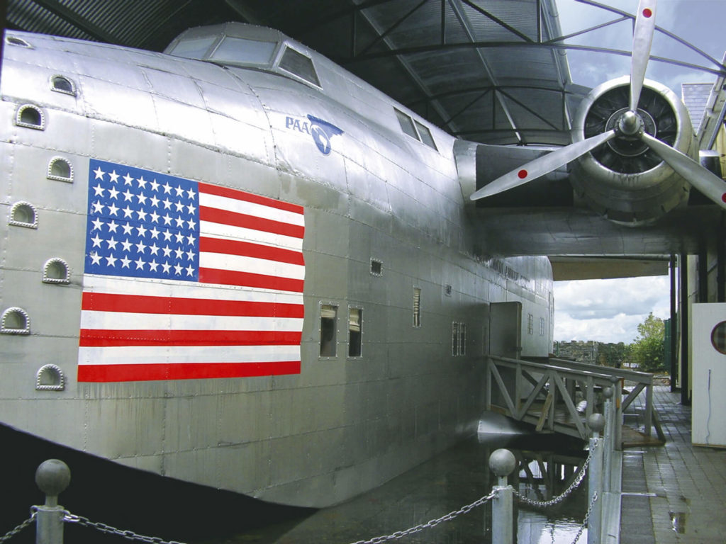 Large silver seaplane with a prominent American flag painted on its side, housed partially under a covered area with one propeller visible.