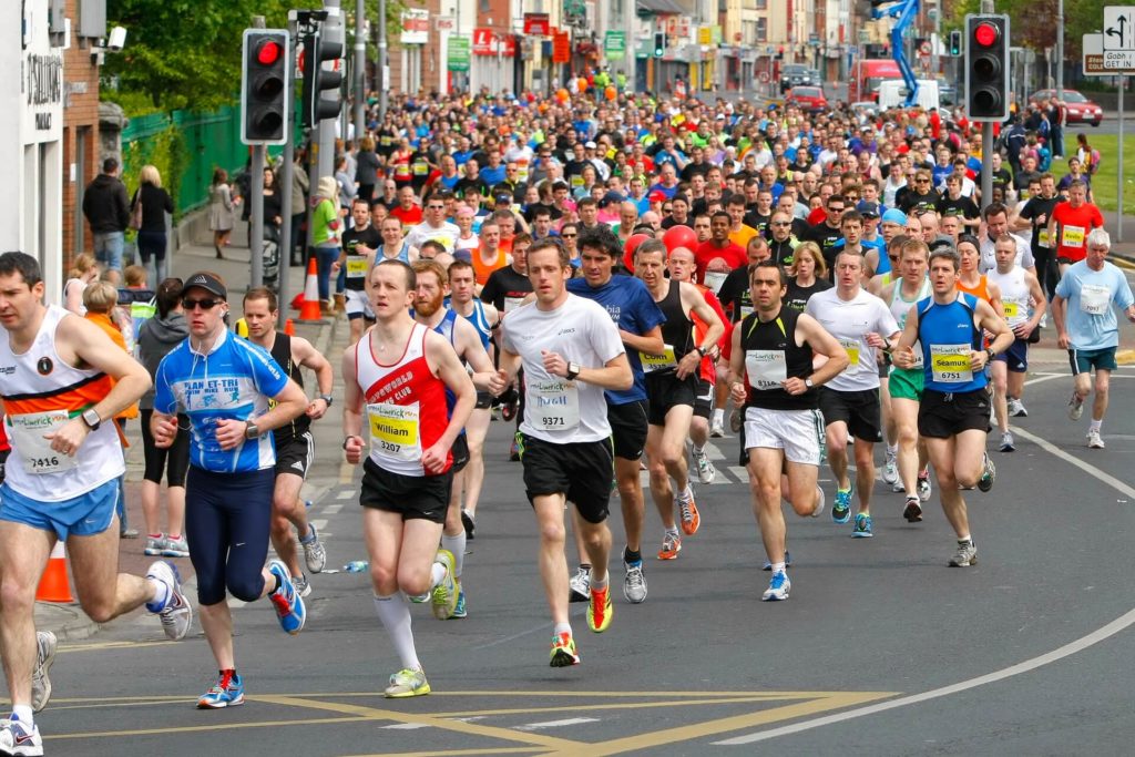 A large group of people runs in a city marathon, wearing race bibs and athletic clothing, with spectators and traffic lights visible in the background.