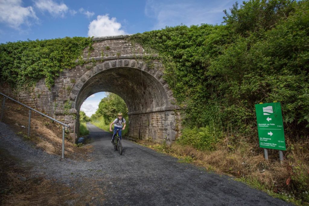 A cyclist rides on a gravel path under a stone arch bridge covered in greenery, with a green direction sign on the right and blue sky overhead.