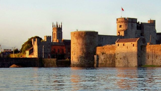 A large stone castle with round towers and a flag on top stands beside a river, with a church and other buildings in the background.
