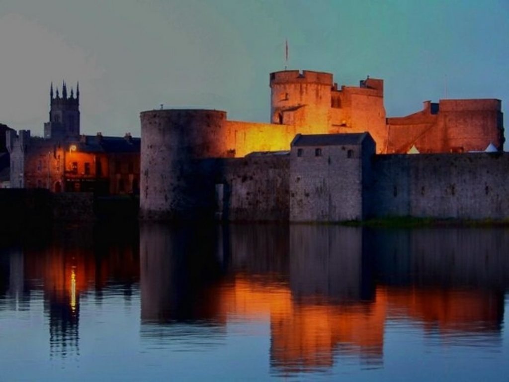 A stone castle with illuminated walls is reflected in calm water at dusk, with a church and other buildings visible in the background.