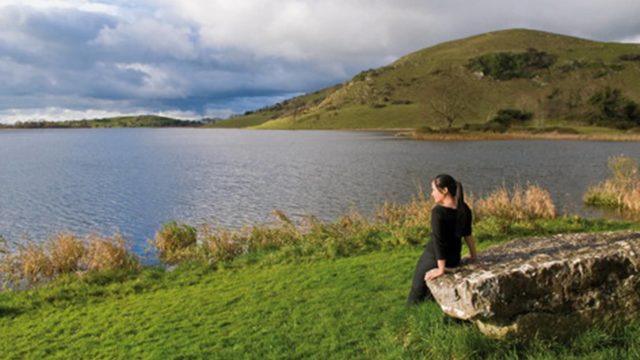 A person in black clothing sits on a large rock beside a lake, with grassy hills and cloudy sky in the background.