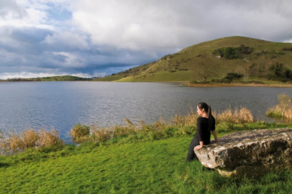 A person in black clothing sits on a stone bench by a grassy lakeshore with hills and cloudy sky in the background.
