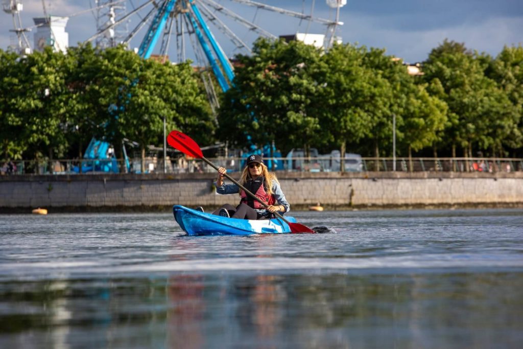 A person paddles a blue kayak on calm water, with trees and a Ferris wheel visible in the background under a partly cloudy sky.