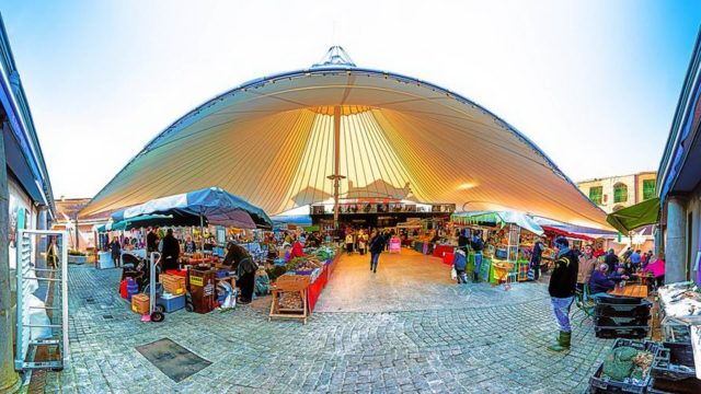An outdoor market scene under a large white canopy with stalls selling various goods and people walking or shopping on a cobblestone surface.