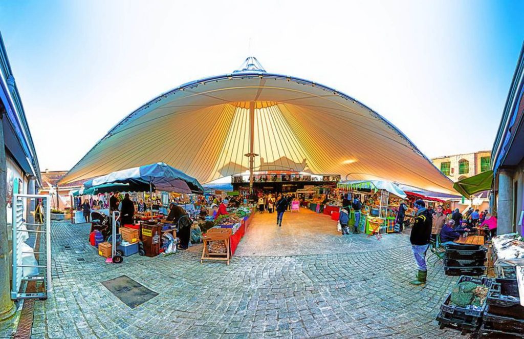 Open-air market scene under a large, white canopy with various stalls selling goods and people browsing the area.