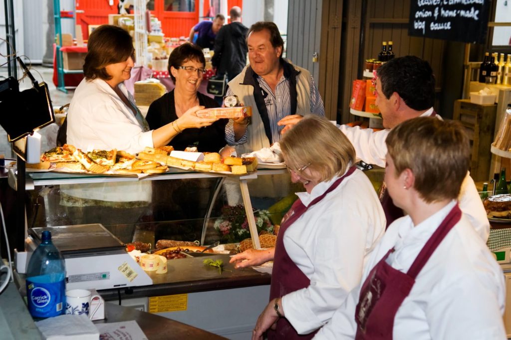 Several people interact at a busy market food counter, with staff serving cheese and baked goods to smiling customers.
