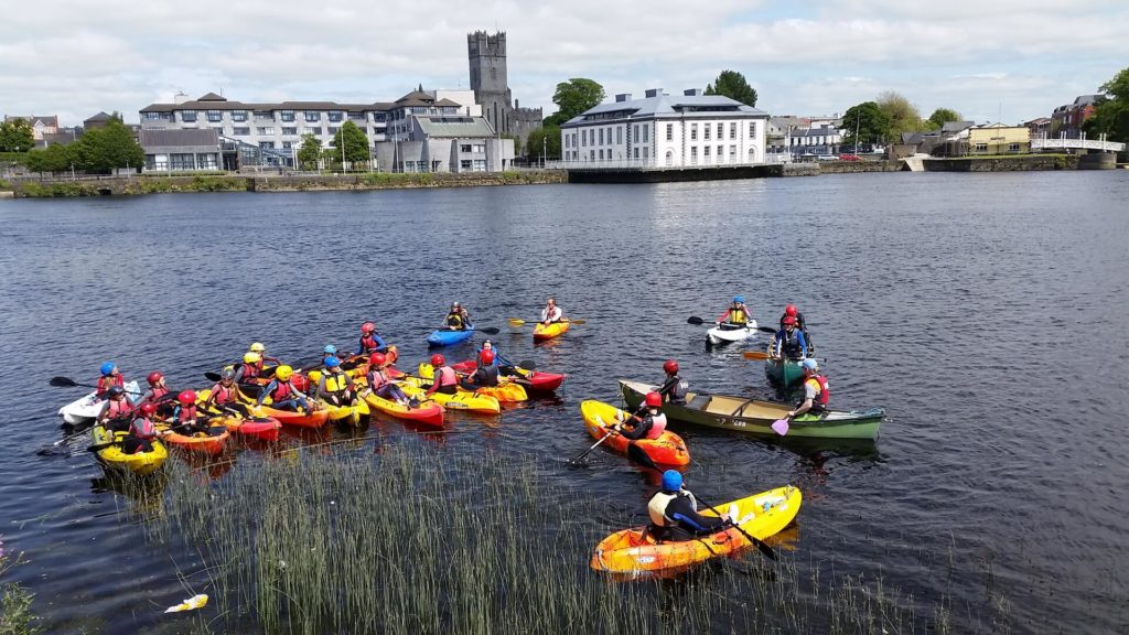 A group of people in colorful kayaks and canoes gather on a river near a city with buildings and a church visible in the background.