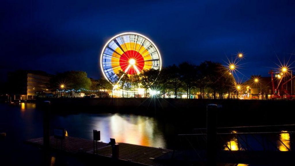A brightly lit Ferris wheel spins at night near a waterfront, with reflections on the water and streetlights illuminating the surrounding area.