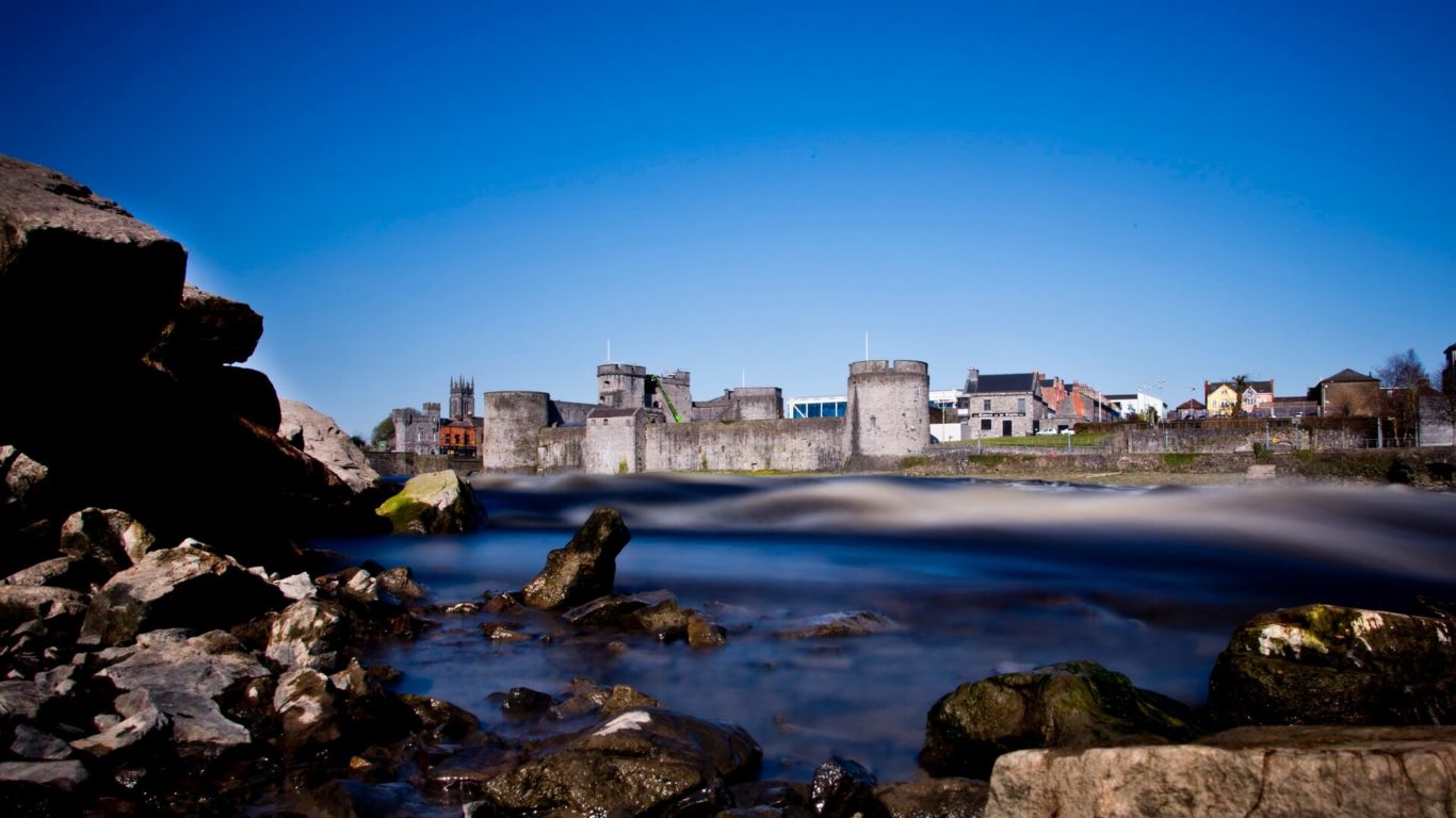 A large stone castle with multiple towers stands beside a river, with rocks and some modern buildings visible under a clear blue sky.