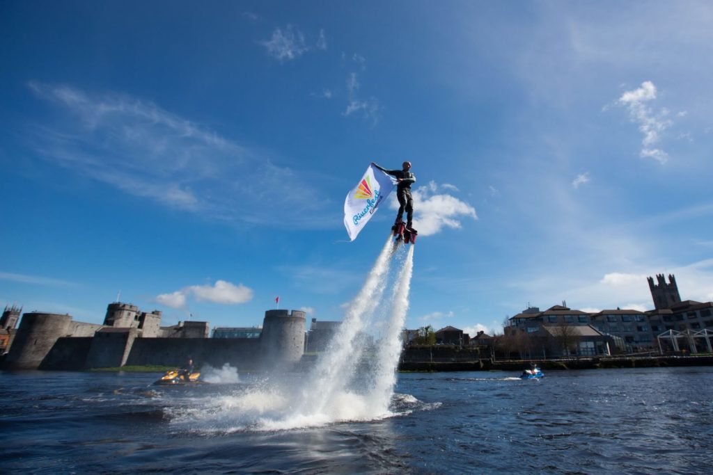 A person uses a water jetpack to rise above a river, holding a flag with colorful graphics; a castle and buildings appear in the background under a blue sky.