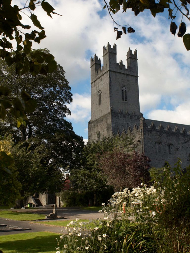 A stone church with a tall, square tower stands amid trees and a garden with white flowers under a partly cloudy sky.