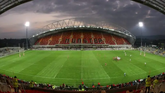 A wide-angle view of a rugby stadium at dusk with teams playing on the field and spectators in the stands, featuring a 