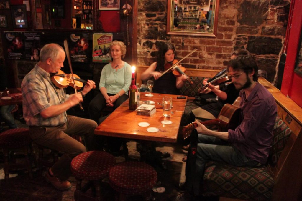 Four people play musical instruments around a wooden table in a cozy pub with stone walls; two others watch and listen.