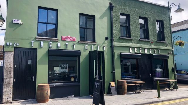 Street view of Treaty City Brewery, a green two-story building with black doors and window frames, outdoor seating, wooden barrels, and a red street sign on the facade.